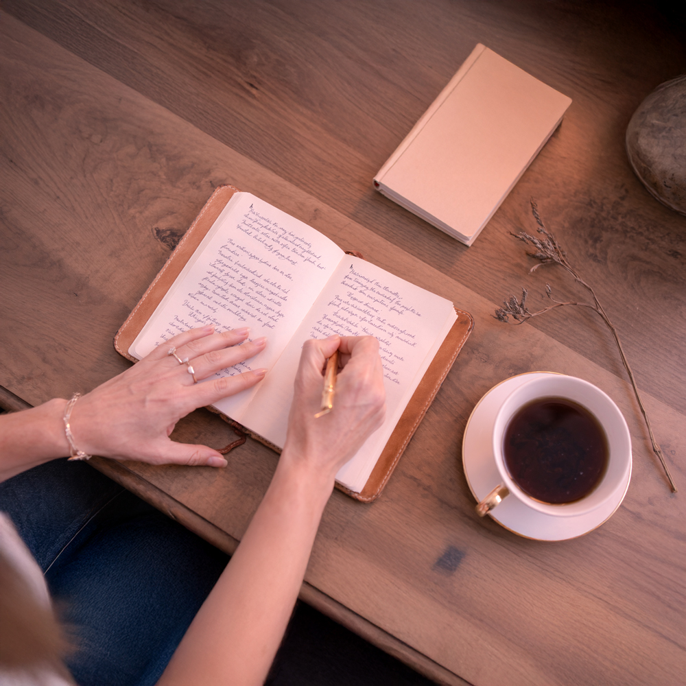 Naomi Zee's hands writing in a camel-leather coaching journal with a brass fountain pen — porcelain teacup beside her on warm walnut, soft rose-gold afternoon light.